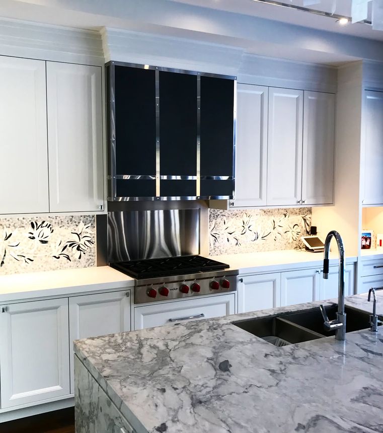 Custom-built range hood and stainless backsplash in black and polished steel, set in a marble-topped kitchen.